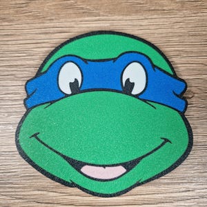 Puede incluir: Primer plano de la cara de una Tortuga Ninja, verde y azul, con una m&aacute;scara azul, ojos blancos y una boca sonriente. La imagen tiene un contorno negro.
