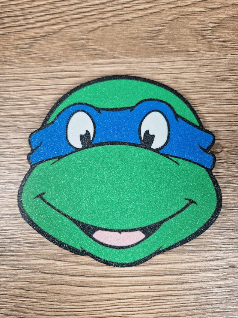 Puede incluir: Primer plano de la cara de una Tortuga Ninja, verde y azul, con una m&aacute;scara azul, ojos blancos y una boca sonriente. La imagen tiene un contorno negro.