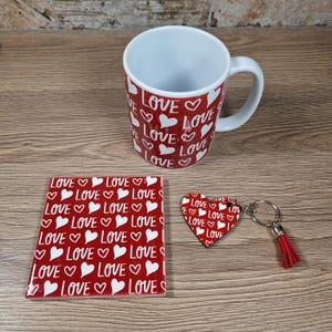 K&ouml;nnte beinhalten: Ein roter Becher, Untersetzer und herzf&ouml;rmiger Schl&uuml;sselanh&auml;nger mit dem Wort "LOVE" und wei&szlig;en Herzen. Der Becher hat einen wei&szlig;en Innenraum und einen Griff. Der Schl&uuml;sselanh&auml;nger hat einen roten Quasten.