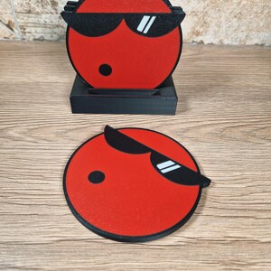 May include: A set of red coasters with a black border and a cartoon face wearing sunglasses. The coasters are round and come with a black holder. The design features a black dot and white accents on the sunglasses.
