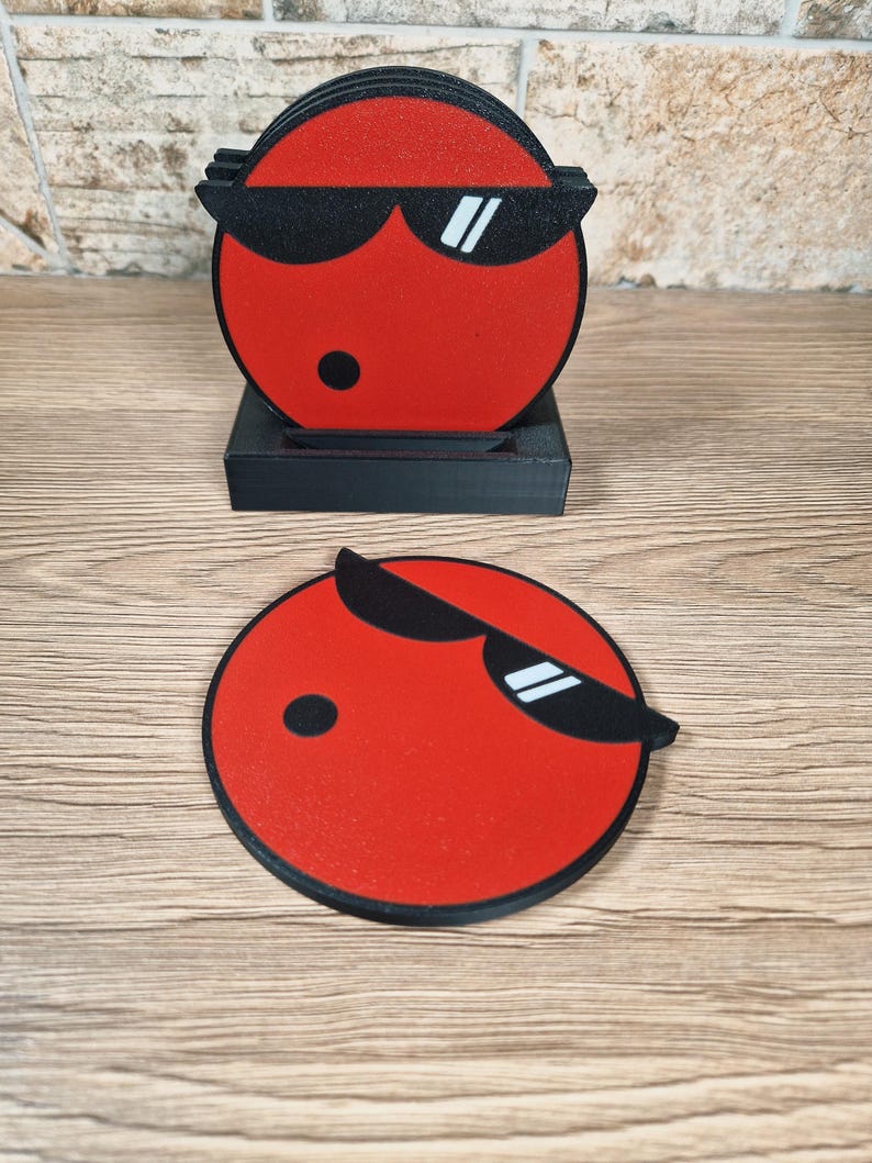 May include: A set of red coasters with a black border and a cartoon face wearing sunglasses. The coasters are round and come with a black holder. The design features a black dot and white accents on the sunglasses.