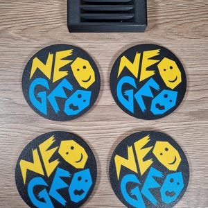 May include: Four black round coasters with the text "NEO GEO" in yellow and blue, along with a smiling face graphic. A black rectangular object with horizontal slits is above the coasters. The coasters are on a wooden surface.