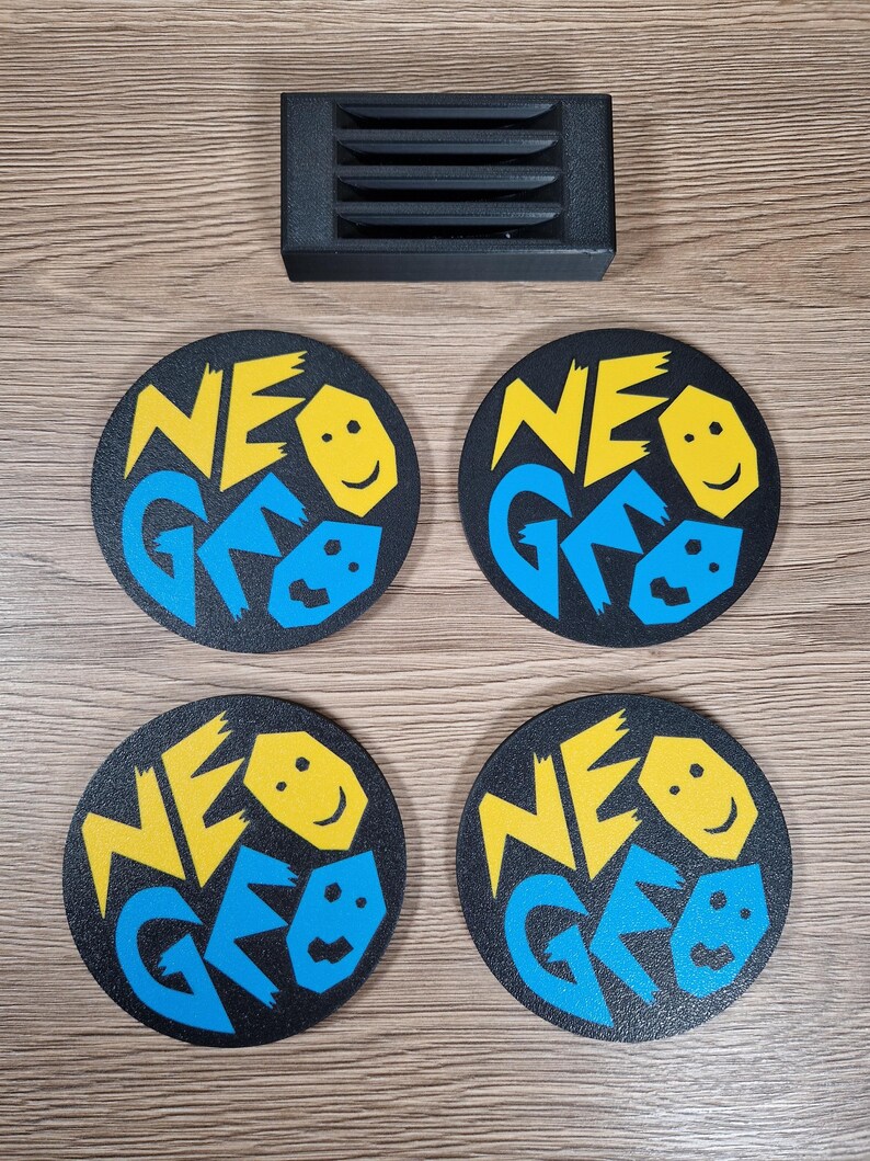 May include: Four black round coasters with the text "NEO GEO" in yellow and blue, along with a smiling face graphic. A black rectangular object with horizontal slits is above the coasters. The coasters are on a wooden surface.