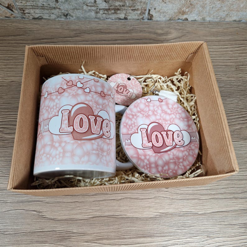 May include: A gift set in a cardboard box, featuring a white mug, a round ornament, and a small tag, all with a pink and white floral pattern and the word "Love". The set includes decorative paper filler.