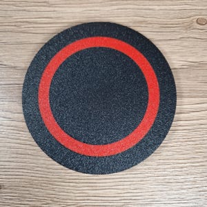 K&ouml;nnte beinhalten: Ein kreisf&ouml;rmiger Untersetzer mit einer strukturierten schwarzen Oberfl&auml;che, die einen markanten, mittig auf der Scheibe platzierten roten Ring aufweist. Das Objekt hat ein flaches Profil und eine gleichm&auml;&szlig;ige matte Oberfl&auml;che.