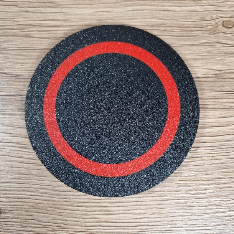 K&ouml;nnte beinhalten: Ein kreisf&ouml;rmiger Untersetzer mit einer strukturierten schwarzen Oberfl&auml;che, die einen markanten, mittig auf der Scheibe platzierten roten Ring aufweist. Das Objekt hat ein flaches Profil und eine gleichm&auml;&szlig;ige matte Oberfl&auml;che.