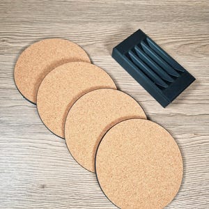 May include: A set of four round cork coasters with black edges, alongside a dark gray holder with slots. The coasters have a natural, light brown color and a textured surface. The holder is rectangular and designed to store the coasters.