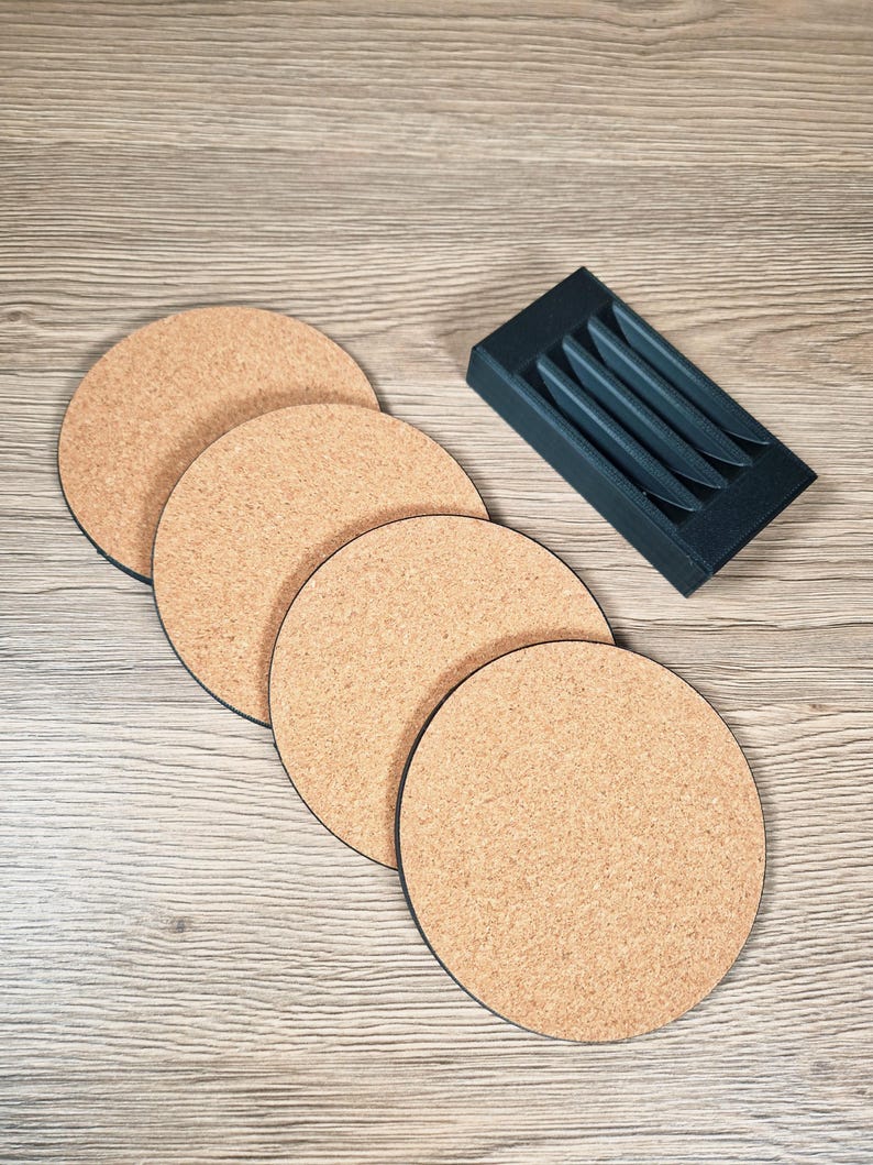 May include: A set of four round cork coasters with black edges, alongside a dark gray holder with slots. The coasters have a natural, light brown color and a textured surface. The holder is rectangular and designed to store the coasters.
