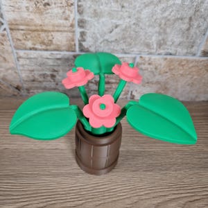 May include: A decorative, 3D-printed flower arrangement featuring pink flowers, green leaves, and a brown pot. The flowers have a layered design, and the leaves are large and heart-shaped. The pot has a barrel-like appearance.