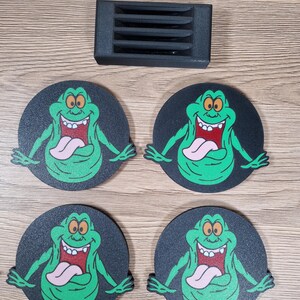 May include: Four black round coasters featuring a green cartoon ghost character with a pink tongue and a wide grin. A black rectangular object with four horizontal slots is also present.