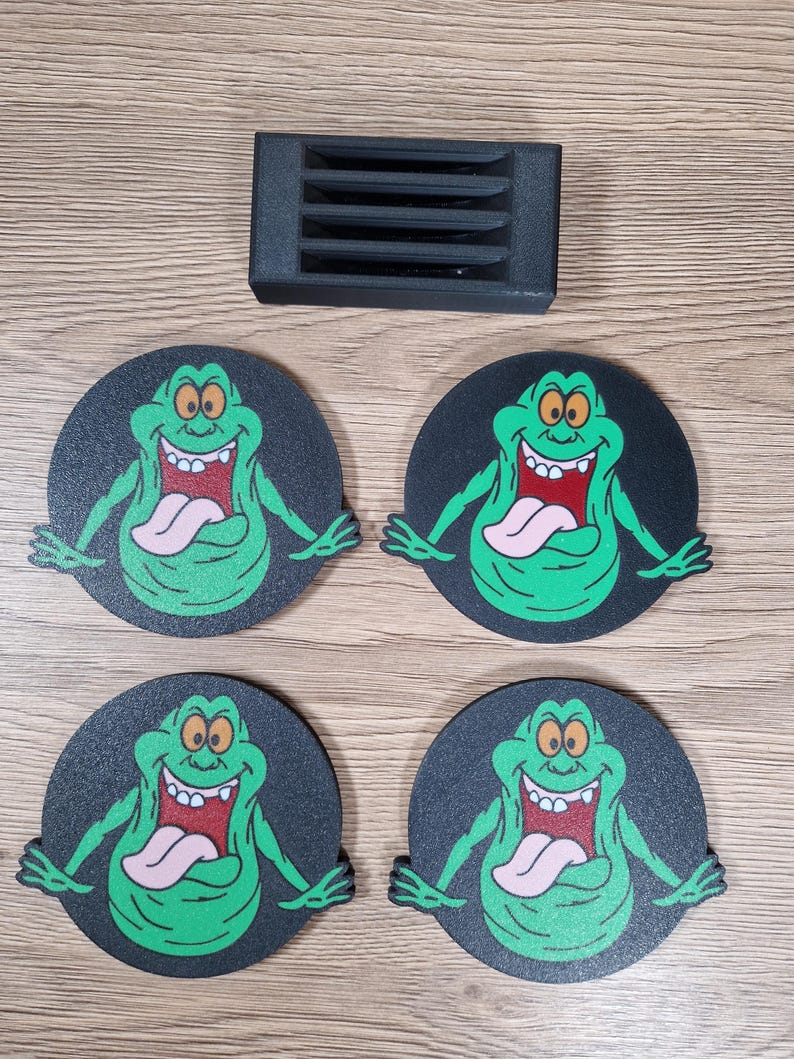 May include: Four black round coasters featuring a green cartoon ghost character with a pink tongue and a wide grin. A black rectangular object with four horizontal slots is also present.