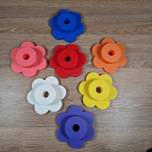 May include: Seven colorful flower-shaped foam objects in shades of pink, red, orange, white, yellow, blue, and purple. Each flower has a circular hole in the center. The objects are arranged on a wooden surface.