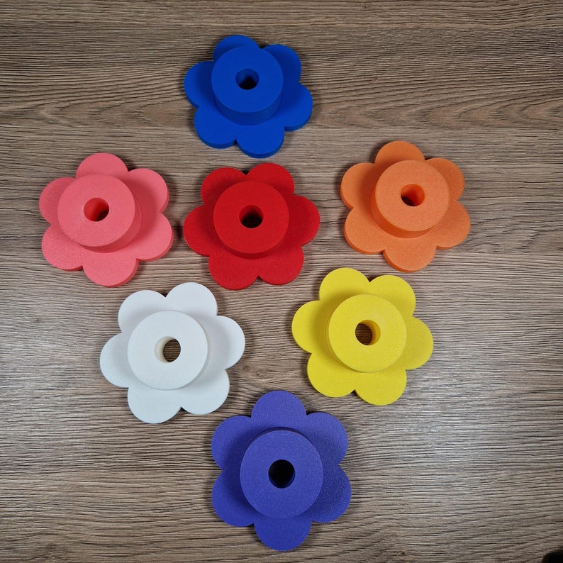 May include: Seven colorful flower-shaped foam objects in shades of pink, red, orange, white, yellow, blue, and purple. Each flower has a circular hole in the center. The objects are arranged on a wooden surface.