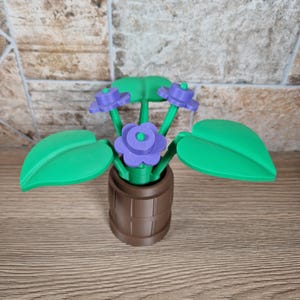 May include: A 3D-printed decorative flower arrangement featuring purple flowers with green centers and leaves. The flowers are in a brown barrel-shaped pot. The leaves are large and green. The arrangement is on a wooden surface.