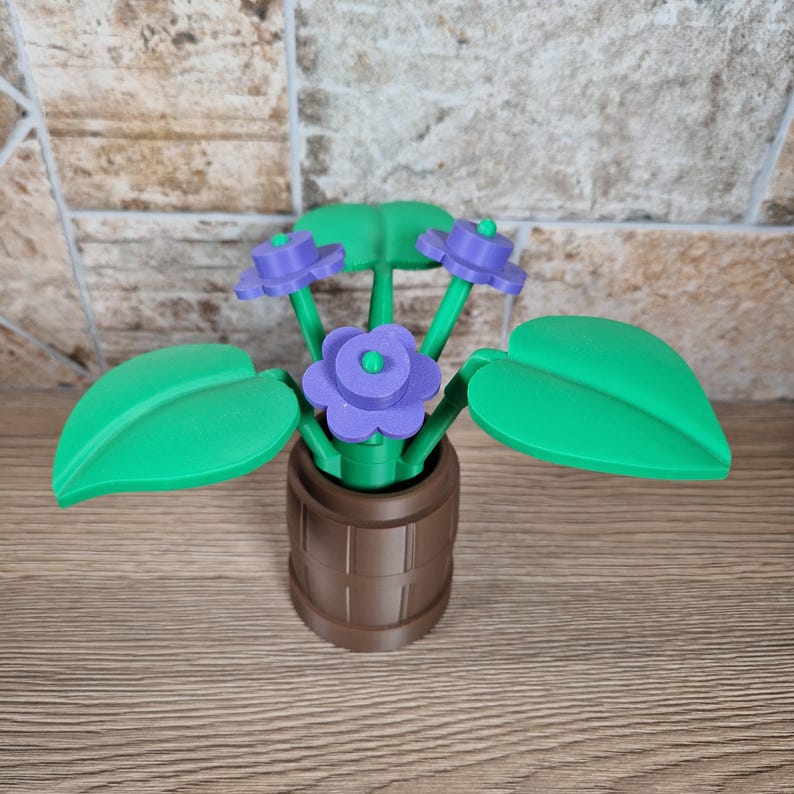 May include: A 3D-printed decorative flower arrangement featuring purple flowers with green centers and leaves. The flowers are in a brown barrel-shaped pot. The leaves are large and green. The arrangement is on a wooden surface.
