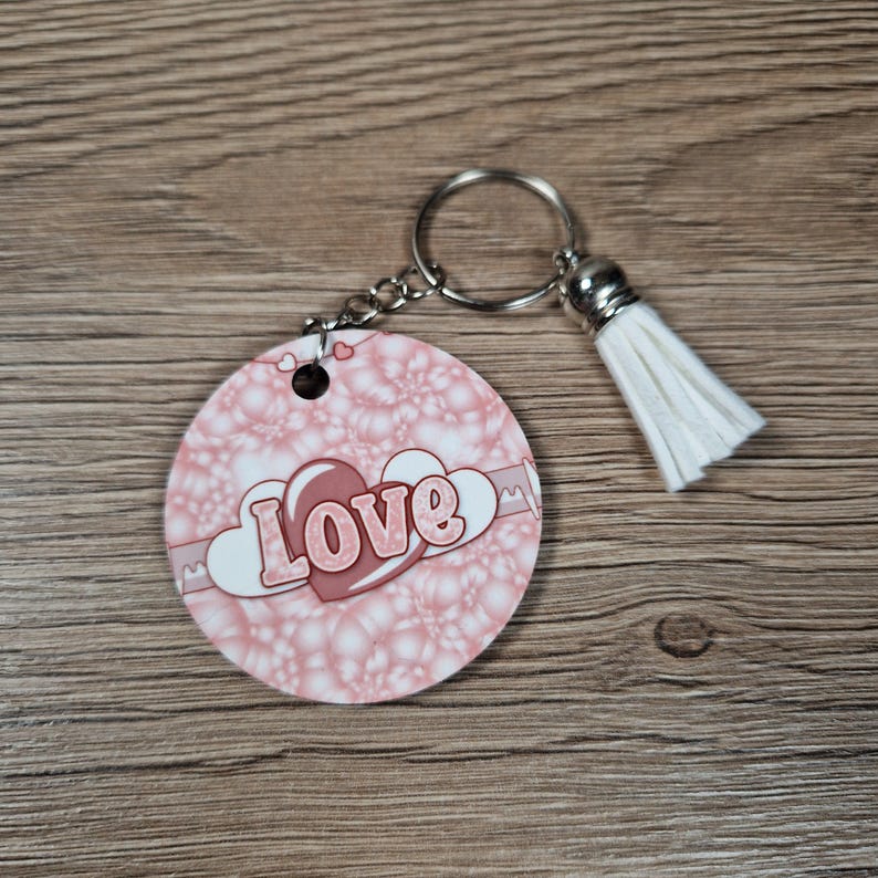 May include: A round keychain with a pink and white bubble pattern. The word "Love" is in the center, with a heart design. A silver chain and keyring are attached, along with a white tassel.