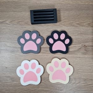 Op de afbeelding: Vier pootvormige onderzetters in grijs, zwart, wit en beige. Elke onderzetter heeft een roze pootafdruk. Een kleine, zwarte rechthoekige zeepbakje met horizontale sleuven is ook aanwezig.