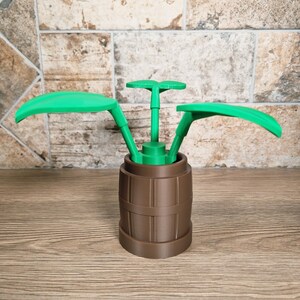 May include: A brown barrel-shaped container with green plant-like elements. The container has a ribbed design, and the green elements include two large, curved leaves and a central stem with a small, flat top. The object is on a wooden surface.