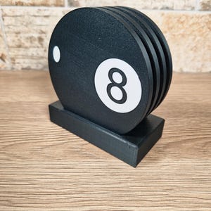 May include: A black wooden coaster set shaped like an eight ball, with a white number eight and a white dot. The coasters are stacked on a black rectangular base, perfect for a game room or bar.