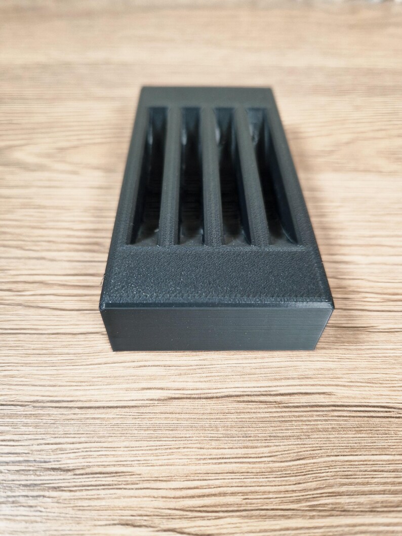 Op de afbeelding: Een rechthoekig, donkergrijs 3D-geprint object met vier verticale sleuven. Het object heeft een getextureerd oppervlak en staat op een lichtbruine houten achtergrond. Het ontwerp suggereert een opslag- of organisatorisch product.