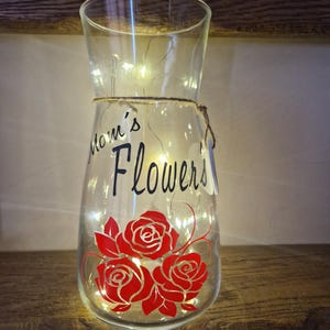 May include: Clear glass vase with a string wrapped around the neck. The vase has a red rose design and the text "Mom's Flowers" on the front. The vase is lit with white fairy lights.