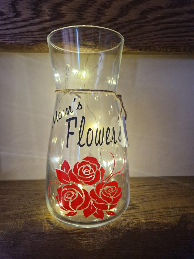 May include: Clear glass vase with a string wrapped around the neck. The vase has a red rose design and the text "Mom's Flowers" on the front. The vase is lit with white fairy lights.