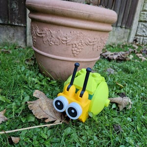 Puede incluir: Un caracol de juguete amarillo y verde con ojos grandes y antenas negras descansa sobre hierba verde. En el fondo, una maceta grande de terracota marr&oacute;n con detalles de vid.