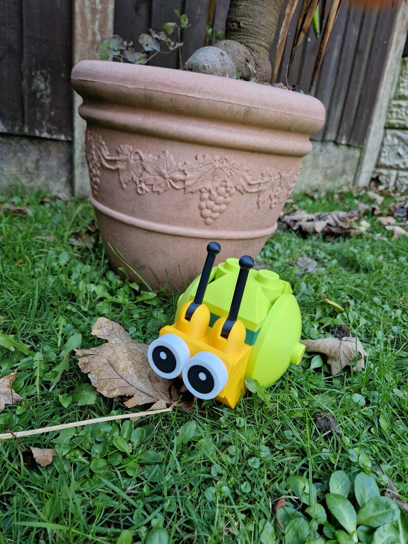 Puede incluir: Un caracol de juguete amarillo y verde con ojos grandes y antenas negras descansa sobre hierba verde. En el fondo, una maceta grande de terracota marr&oacute;n con detalles de vid.