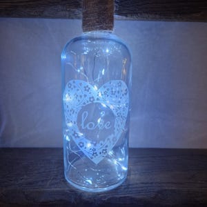 May include: A clear glass bottle with a burlap wrapped top and a white heart design with the word "love" inside. The bottle is filled with white fairy lights.
