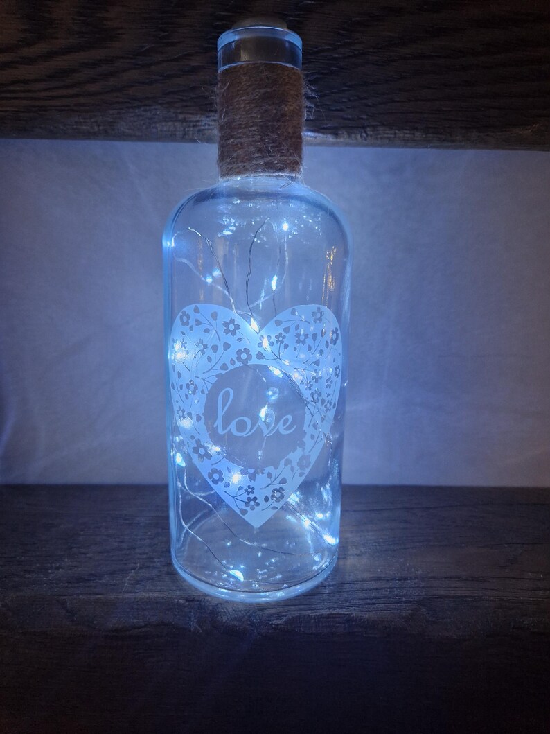 May include: A clear glass bottle with a burlap wrapped top and a white heart design with the word "love" inside. The bottle is filled with white fairy lights.