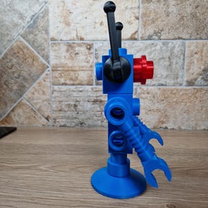 May include: A blue, red, and black robot toy with a round base and articulated arms. The robot has a gear-like component and two black levers extending upwards. The toy is made of plastic and stands upright on a wooden surface.