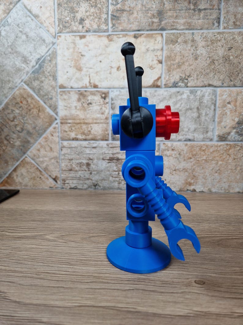 May include: A blue, red, and black robot toy with a round base and articulated arms. The robot has a gear-like component and two black levers extending upwards. The toy is made of plastic and stands upright on a wooden surface.