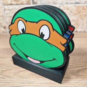 Puede incluir: Un conjunto de posavasos de las Tortugas Ninja. Cada posavasos es verde con una m&aacute;scara naranja, ojos blancos y una boca sonriente. Los posavasos est&aacute;n apilados en un soporte rectangular negro.