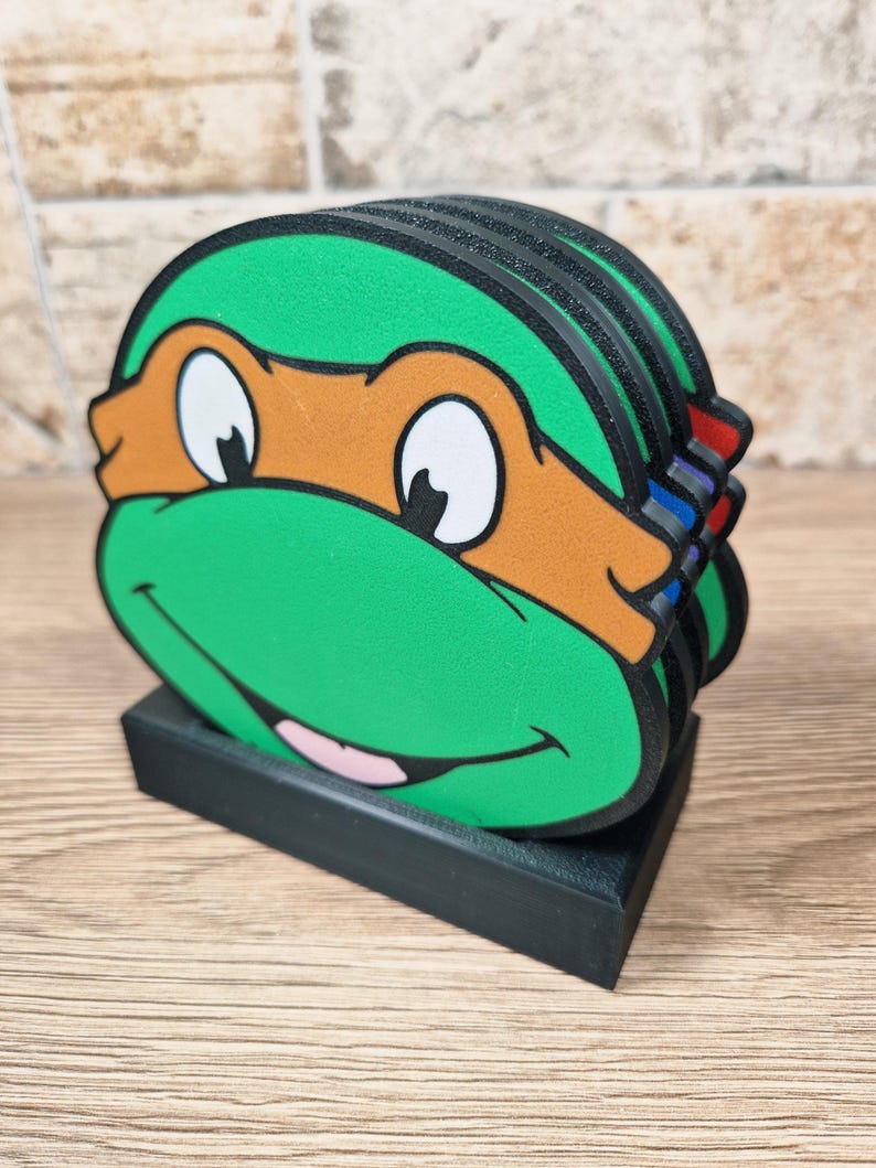 Puede incluir: Un conjunto de posavasos de las Tortugas Ninja. Cada posavasos es verde con una m&aacute;scara naranja, ojos blancos y una boca sonriente. Los posavasos est&aacute;n apilados en un soporte rectangular negro.