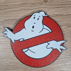 K&ouml;nnte beinhalten: Rundes, rot-schwarzes Ghostbusters-Logo mit einer wei&szlig;en Geisterfigur, die die Mitte durchkreuzt. Der Geist hat einen &uuml;berraschten Gesichtsausdruck. Das Logo befindet sich auf einer Holzoberfl&auml;che.
