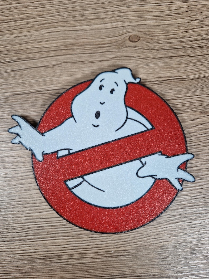 K&ouml;nnte beinhalten: Rundes, rot-schwarzes Ghostbusters-Logo mit einer wei&szlig;en Geisterfigur, die die Mitte durchkreuzt. Der Geist hat einen &uuml;berraschten Gesichtsausdruck. Das Logo befindet sich auf einer Holzoberfl&auml;che.