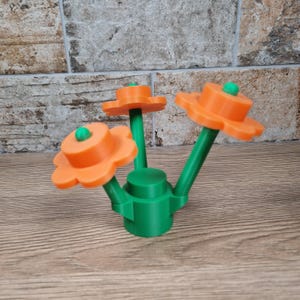 May include: A 3D-printed decorative flower arrangement. The arrangement features three orange flower heads with green stems and a green base. Each flower has a small green sphere in the center. The object is set against a wood-grain surface.