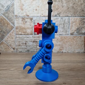 May include: A blue robot toy with a red head and black details. The robot has a claw arm and a round base. The robot is standing on a wooden surface.