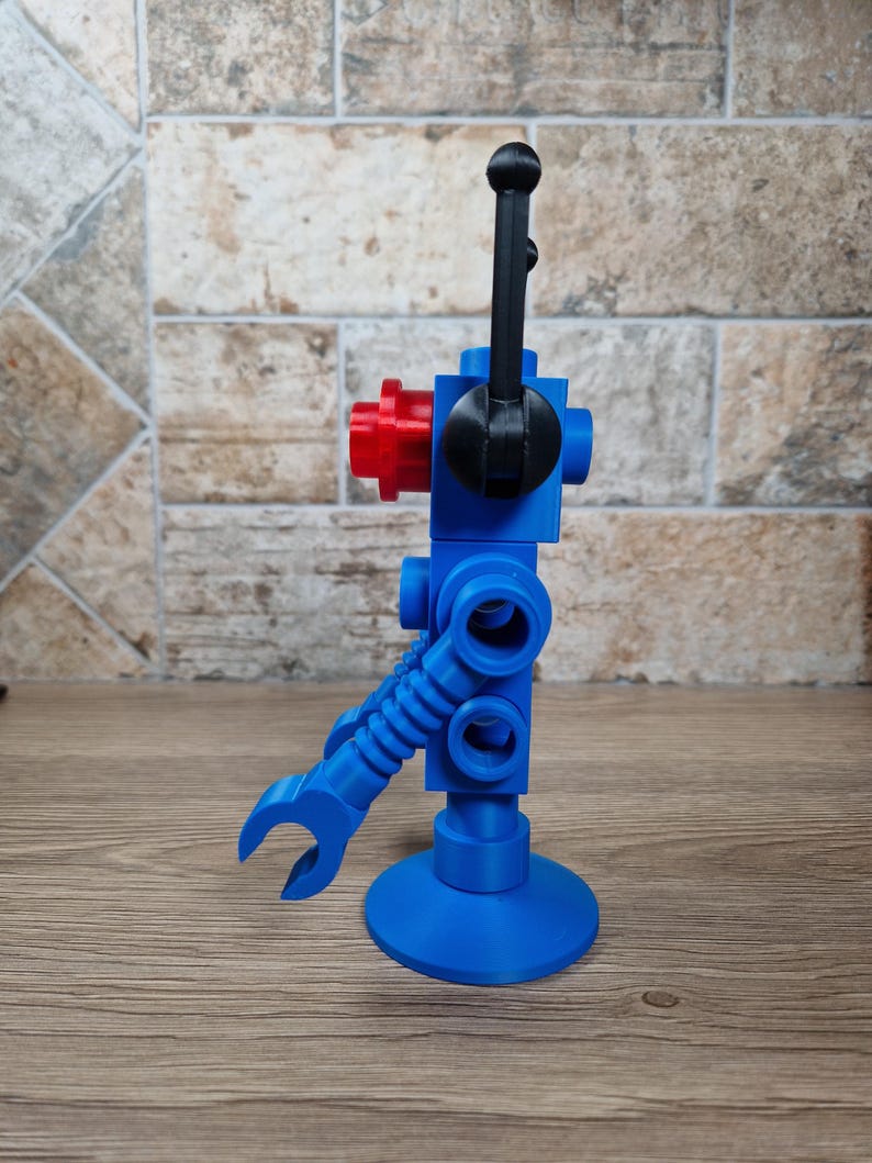 May include: A blue robot toy with a red head and black details. The robot has a claw arm and a round base. The robot is standing on a wooden surface.