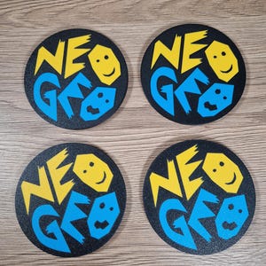 May include: Four black coasters with the text "NEO GEO" in yellow and blue. Each coaster features a smiling face graphic. The coasters are round and appear to be made of a textured material.