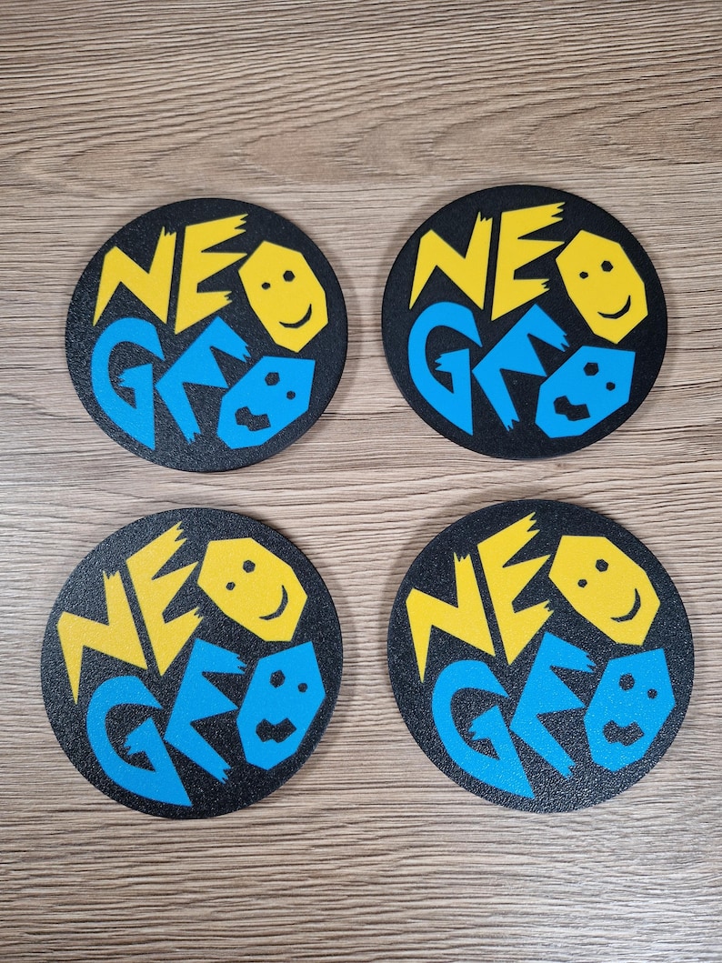 May include: Four black coasters with the text "NEO GEO" in yellow and blue. Each coaster features a smiling face graphic. The coasters are round and appear to be made of a textured material.