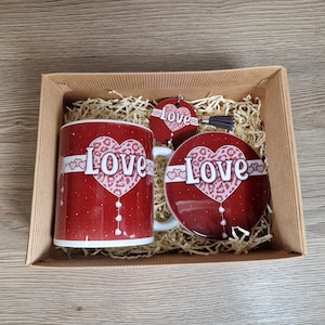 May include: A gift set in a cardboard box, featuring a red mug, coaster, and keychain. Each item has a heart design with the word "Love" and a leopard print pattern. The mug is white with a handle.