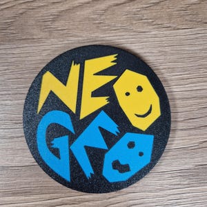 May include: A black, round coaster with the words "NEO GEO" in yellow and blue, along with a smiling face graphic. The coaster is likely made of a durable material and is designed to protect surfaces from drink condensation.