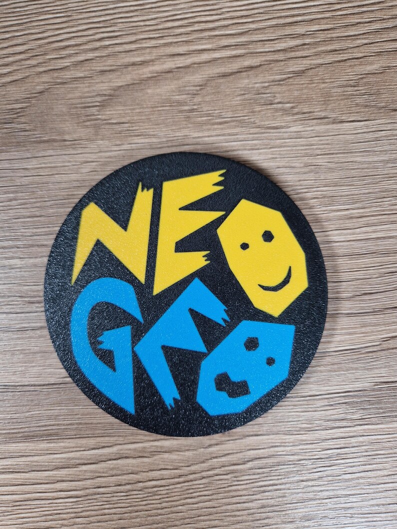 May include: A black, round coaster with the words "NEO GEO" in yellow and blue, along with a smiling face graphic. The coaster is likely made of a durable material and is designed to protect surfaces from drink condensation.