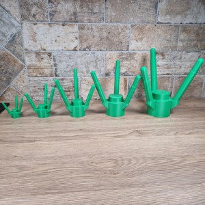 May include: A collection of five green watering can attachments, varying in size, arranged in a row. Each attachment has a cylindrical base with a spout and multiple angled arms. The items are displayed on a wooden surface.
