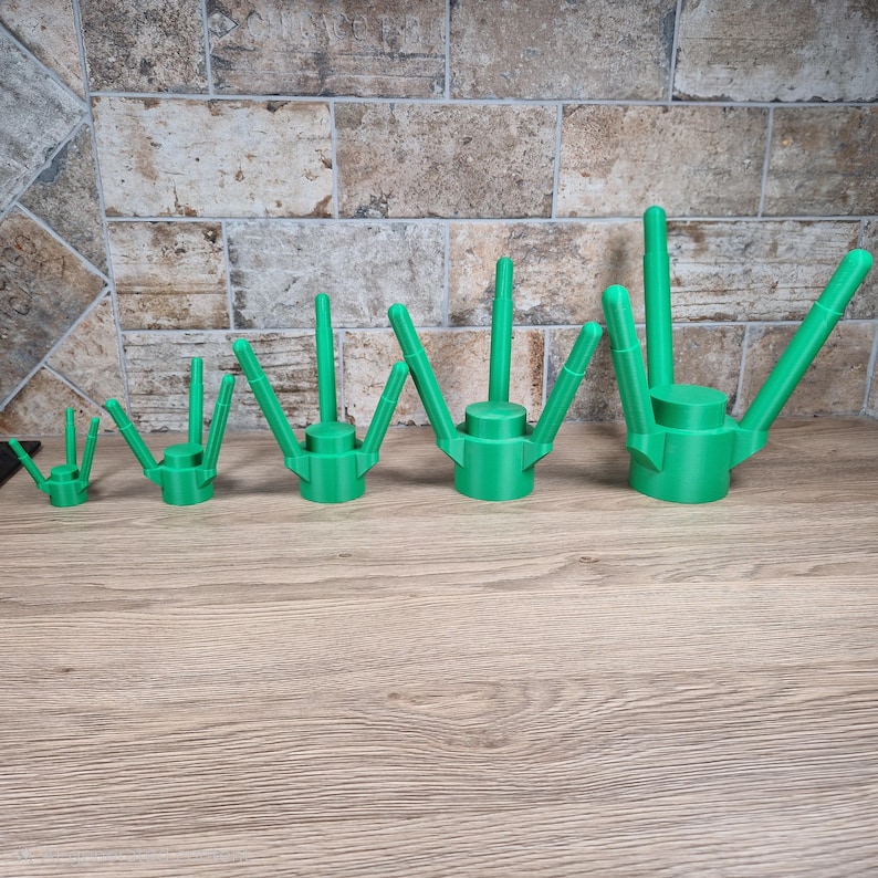 May include: A collection of five green watering can attachments, varying in size, arranged in a row. Each attachment has a cylindrical base with a spout and multiple angled arms. The items are displayed on a wooden surface.