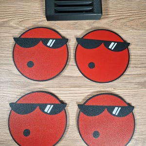 May include: Four red circular coasters with black sunglasses and a black dot, arranged with a black rectangular object with horizontal slits. The coasters have a textured surface and a black outline. The items are on a wooden surface.