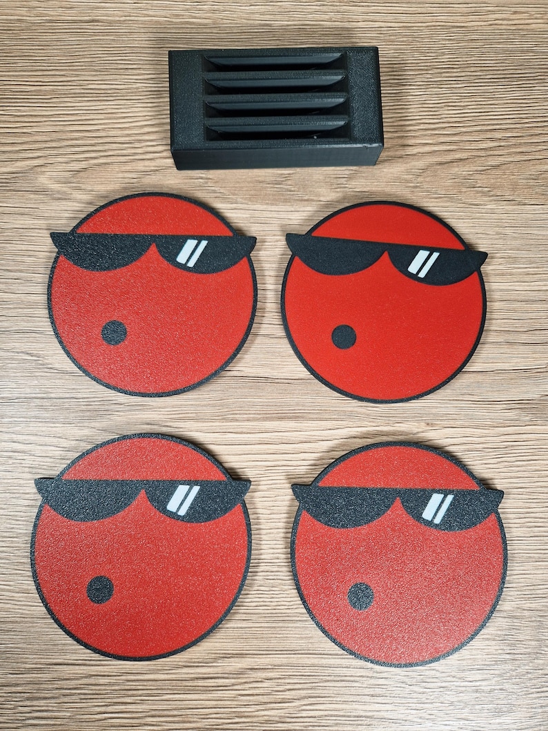 May include: Four red circular coasters with black sunglasses and a black dot, arranged with a black rectangular object with horizontal slits. The coasters have a textured surface and a black outline. The items are on a wooden surface.