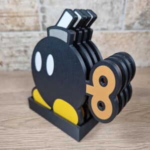 May include: A set of black and yellow Bomb-Omb coasters, a character from the Super Mario video game series. The coasters have a black base and a wind-up key design. The set includes multiple coasters, perfect for protecting surfaces from drink rings.