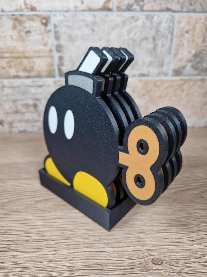 May include: A set of black and yellow Bomb-Omb coasters, a character from the Super Mario video game series. The coasters have a black base and a wind-up key design. The set includes multiple coasters, perfect for protecting surfaces from drink rings.